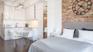 A modern hotel room with a kitchen area, a dining table with chairs, a large bed, and a wall clock.