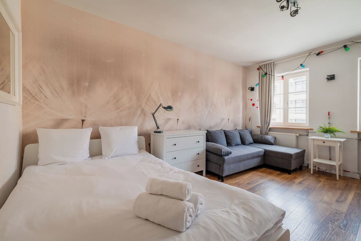 Un dormitorio con una cama grande, un sofá gris, una mesita de noche y una ventana con cortinas.