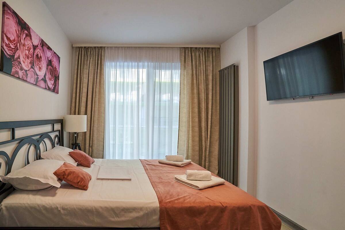 A hotel room with a bed, a TV, a painting, and a window with curtains.