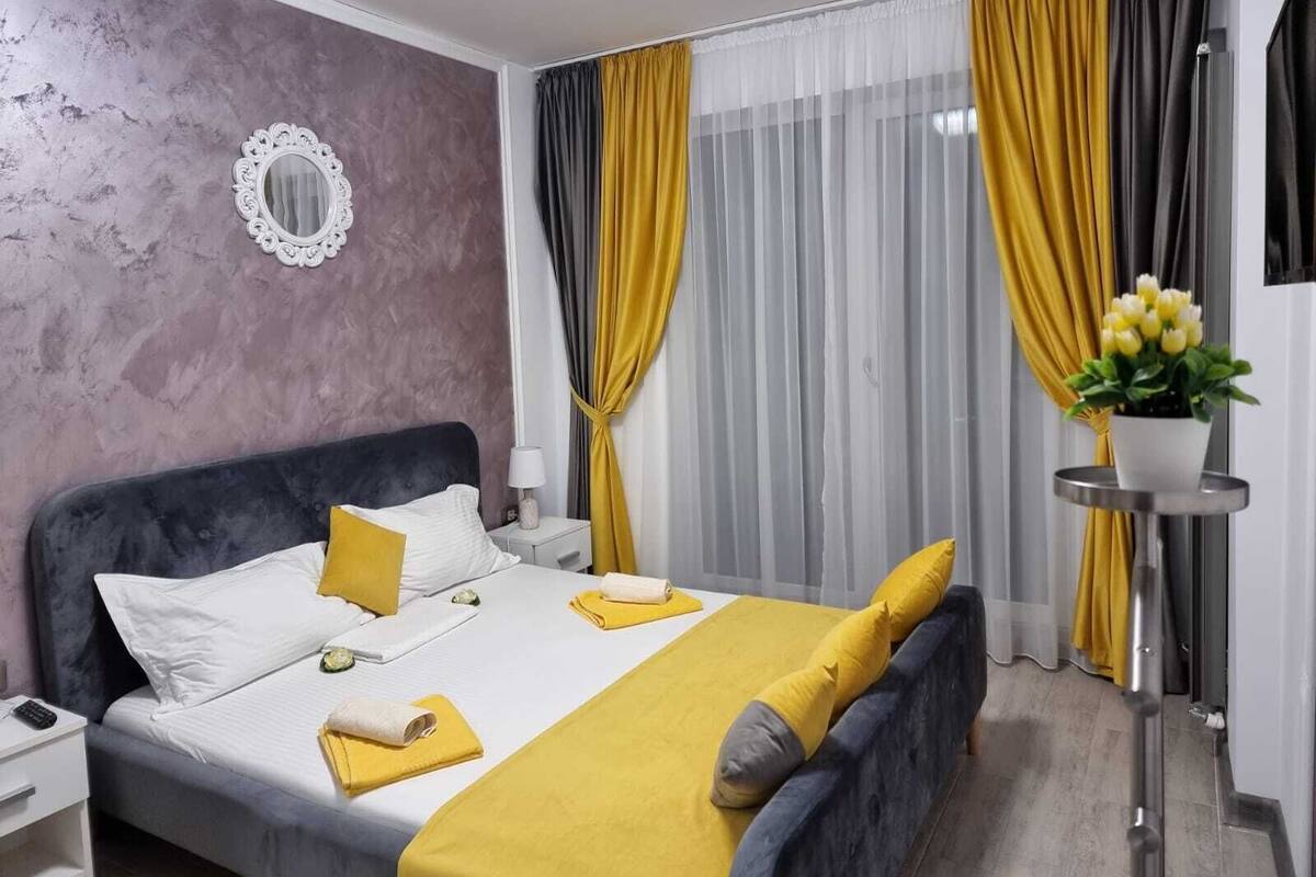 A modern hotel room with a large bed, a bedside table, a mirror, and a small side table with a lamp and a potted plant.