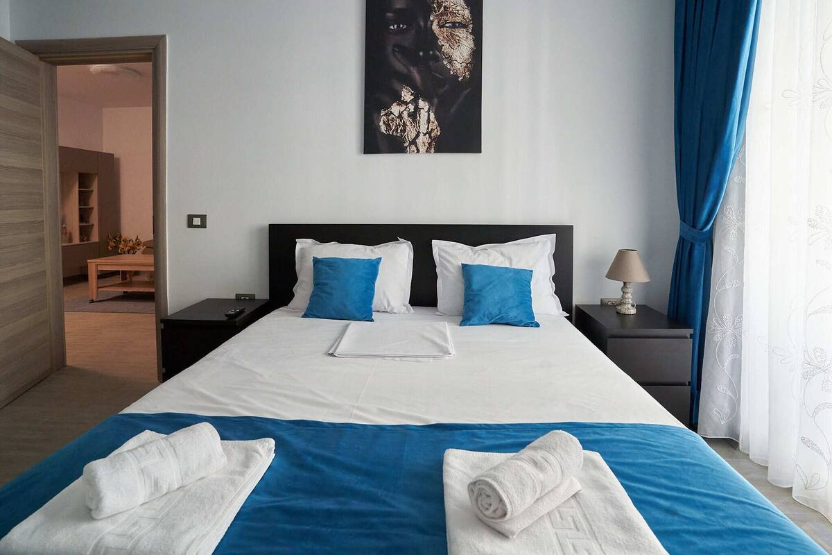 A neatly made bed with blue and white bedding, a bedside table with a lamp, and a painting on the wall.