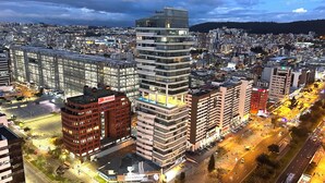 Paisagem urbana com edifícios modernos, um imponente arranha-céu de vidro e um cruzamento bem iluminado.