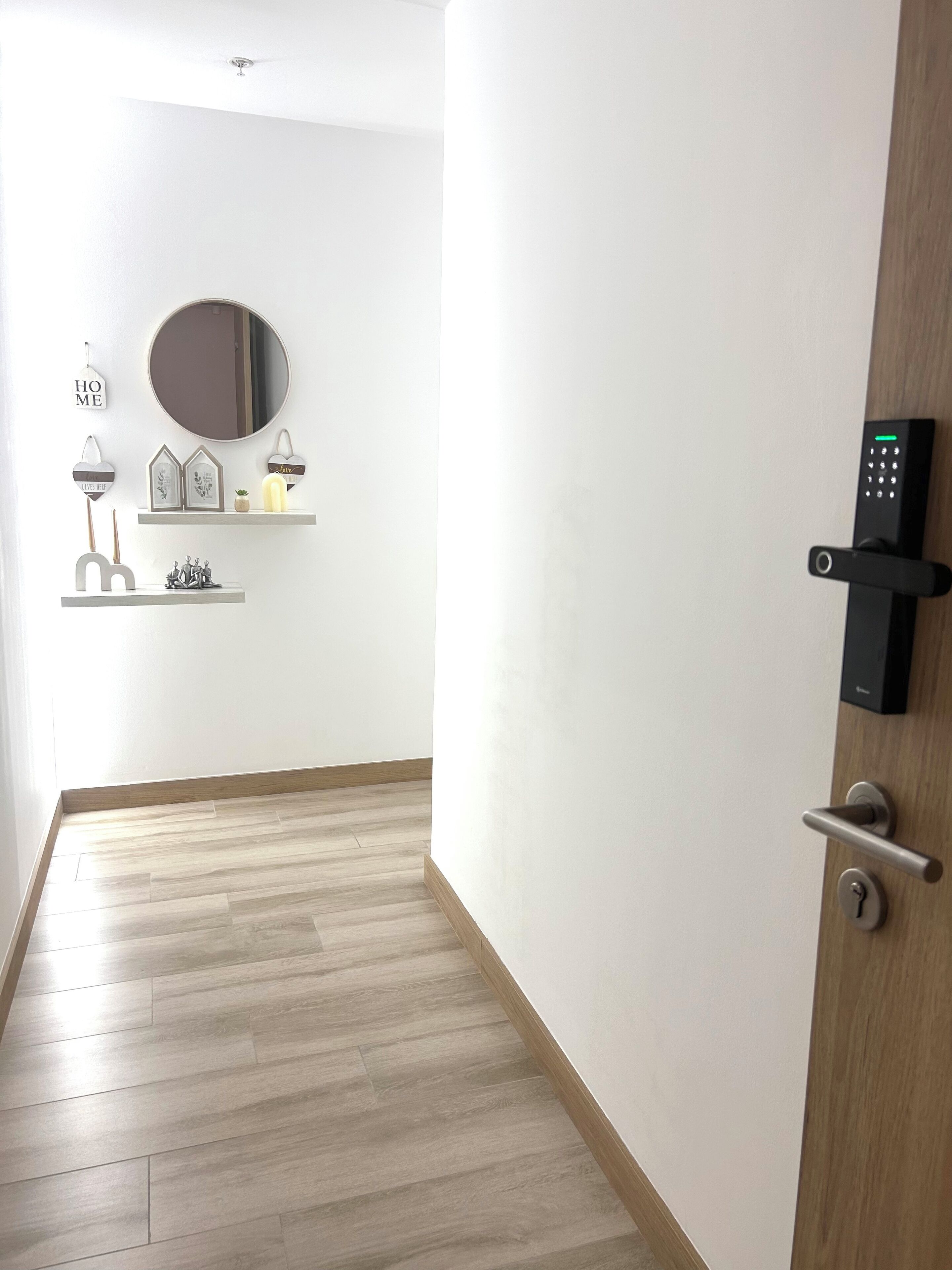 A hallway with a wooden floor, a door with a keypad lock, and wall-mounted shelves with decorative items.