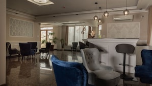Een ruime lobby met een gepolijste marmeren vloer, diverse zitmogelijkheden waaronder een blauwe velours fauteuil, een grijze gestoffeerde fauteuil en een barkruk. Er hangen meerdere pendelverlichting aan het plafond, en een airconditioner is aan de muur bevestigd.