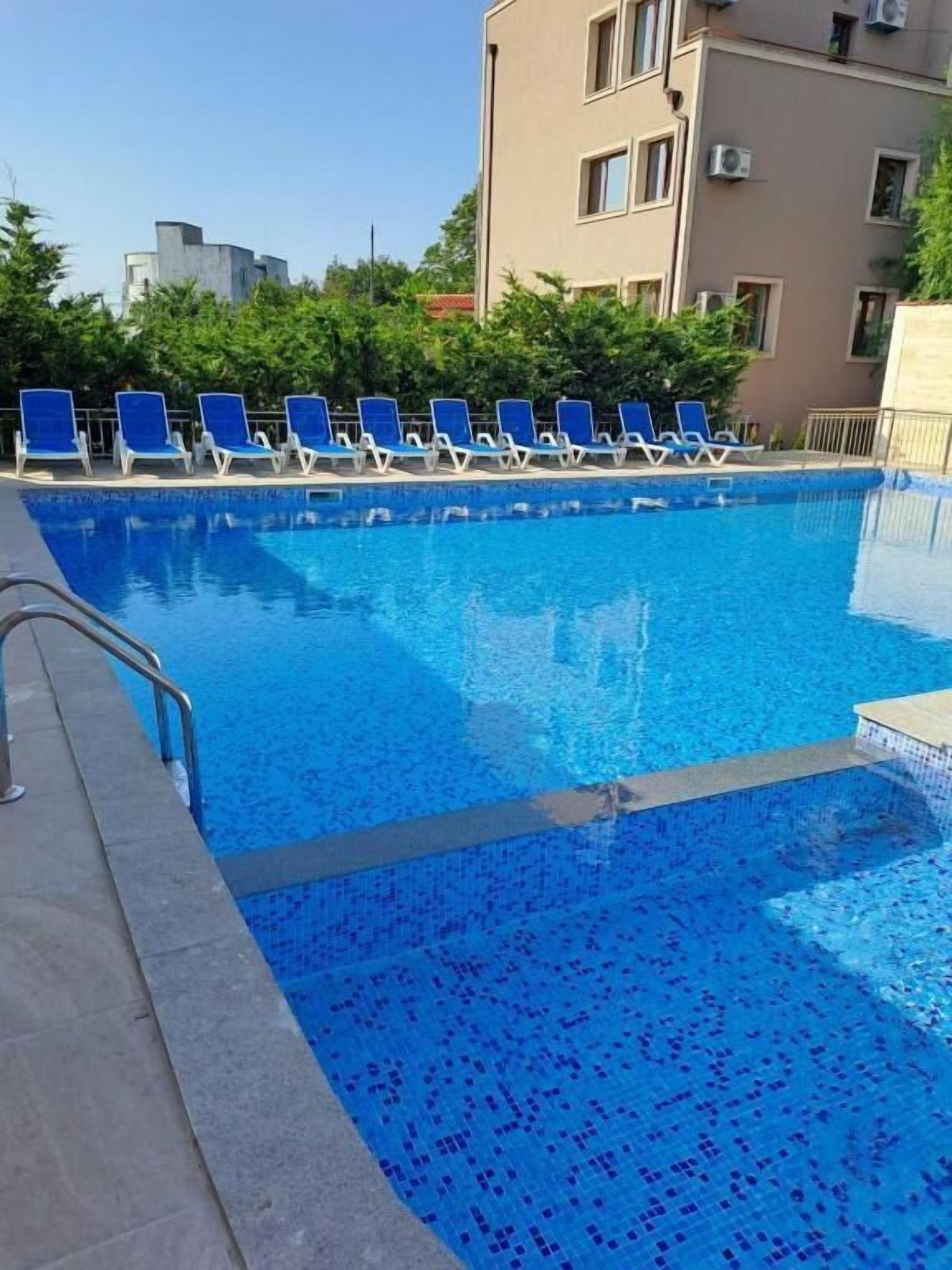 Piscina con azulejos azules, rodeada de sillones de descanso y un edificio al fondo.