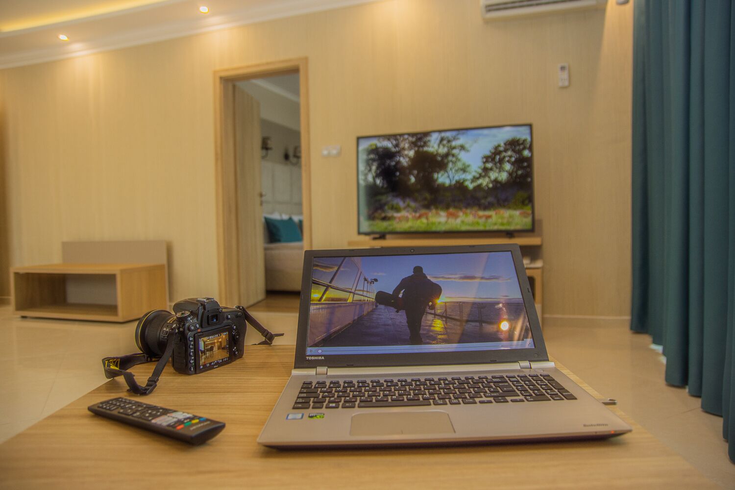 Un ordinateur portable est posé sur une table en bois, avec un appareil photo et une télécommande à côté. On aperçoit une télévision à l’arrière-plan, et une porte donne accès à une autre pièce.