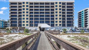A multi-story hotel with a central entrance, a covered walkway, and a parking area with cars. - HEART OF GULF SHORES, GULF-FRONT BALCONY, POOL! | BEACHBALL PROPERTIES (Gulf Shores)