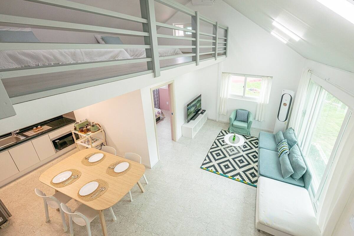 A compact, modern apartment with a loft bedroom, a dining area, a living room with a sofa and TV, and a kitchen with a sink and counter.