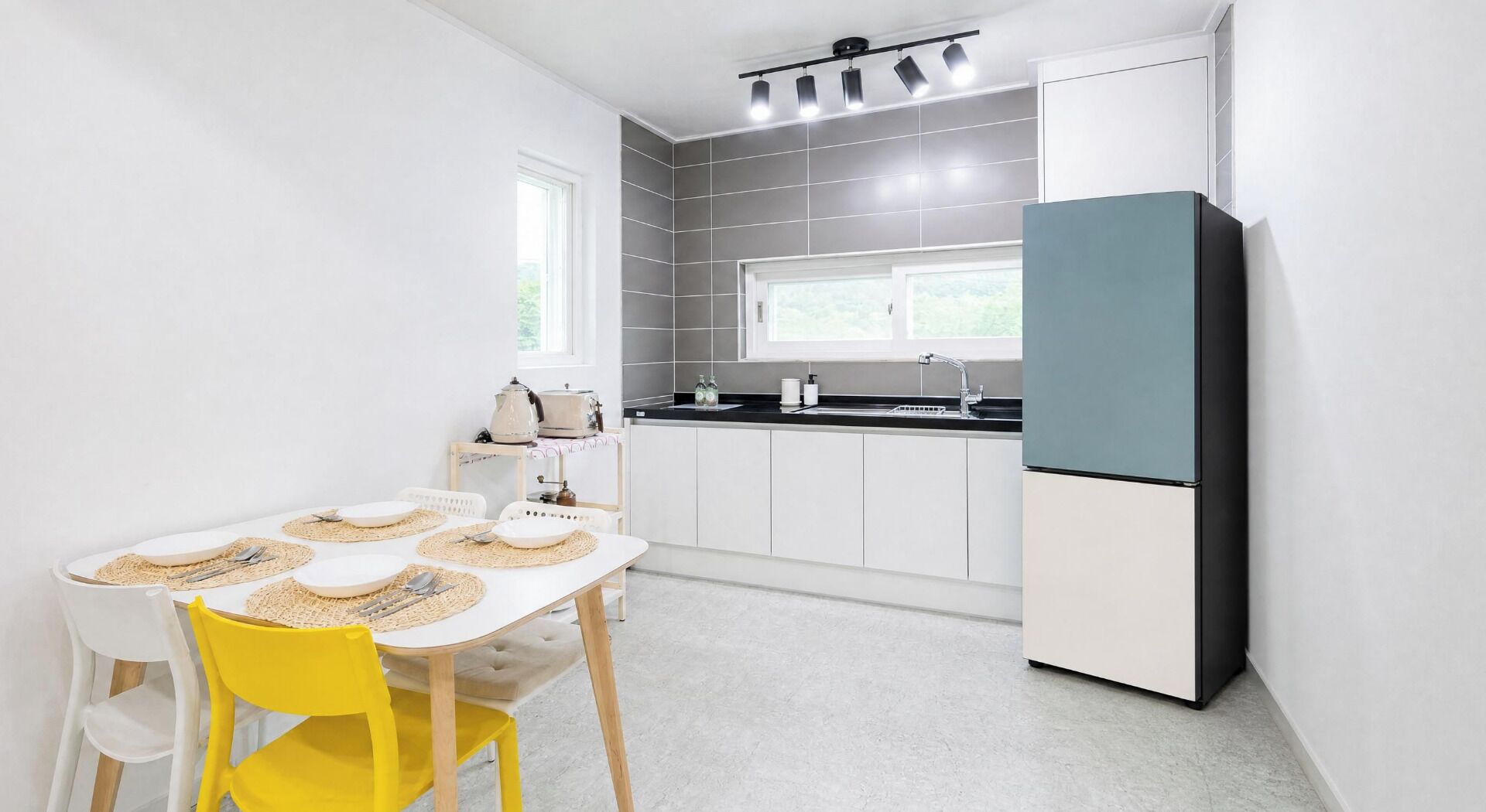 A compact kitchen with white cabinets, a black refrigerator, and a dining area with a white table and yellow chairs.