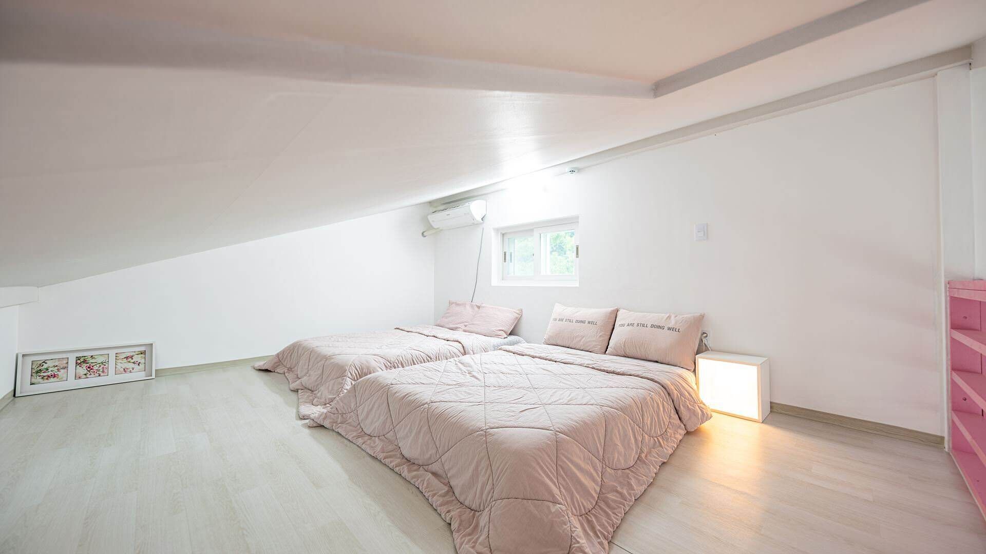 Un dormitorio con techo inclinado, una ventana, una cama con dos almohadas y una mesita de noche con una lámpara.