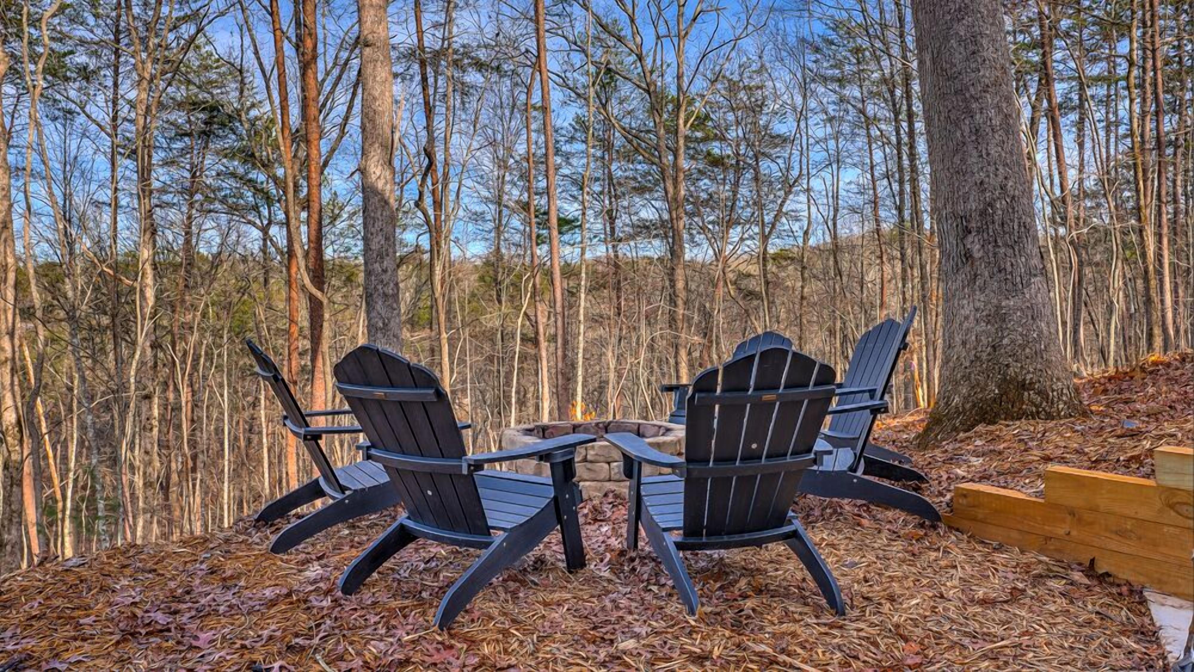 Two Adirondack chairs facing each other around a stone fire pit in a forest setting.