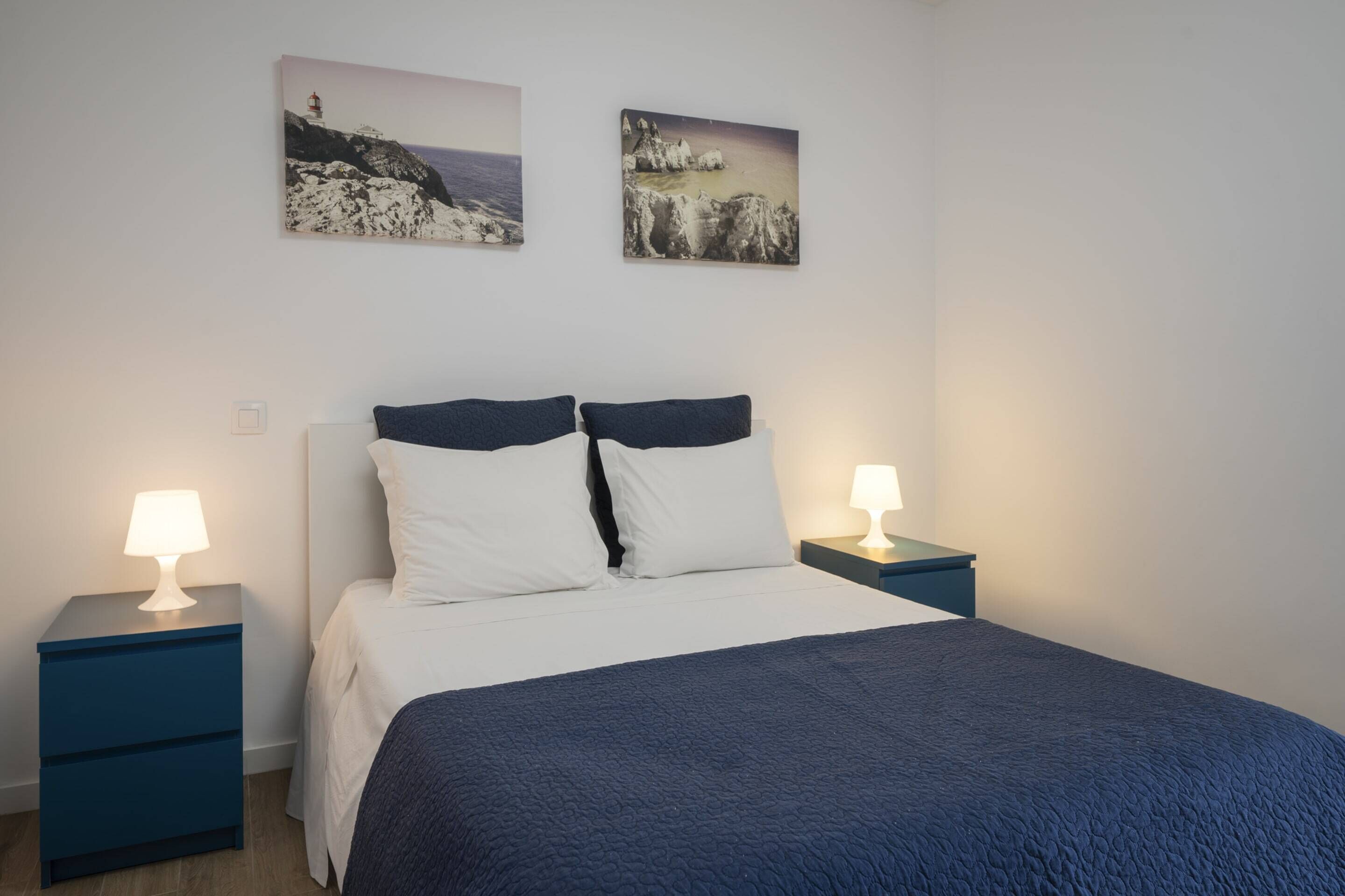 Un dormitorio con una cama, dos mesitas de noche, dos lámparas de pared y dos cuadros con escenas costeras.