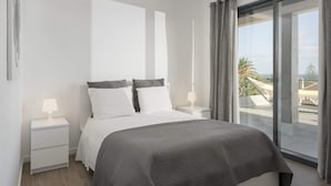 A modern bedroom with a large bed, white nightstands, and a view of a balcony with outdoor furniture. - In the Heart of Porto Santo - Casa da Maresia (Porto Santo)