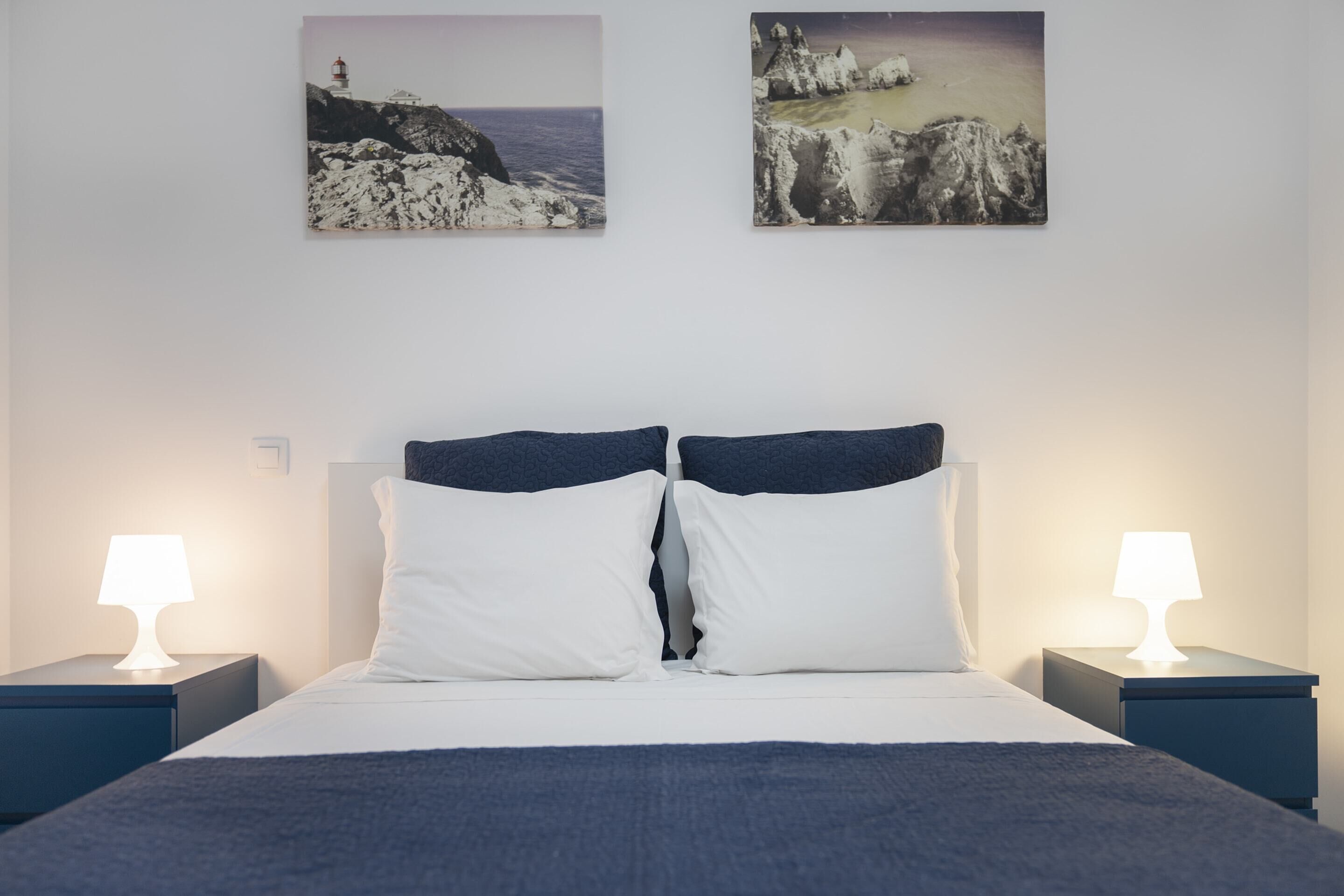 A neatly made bed with white and navy bedding, flanked by two bedside tables with lamps, and two framed coastal landscape paintings on the wall.