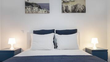 A neatly made bed with white and navy bedding, flanked by two bedside tables with lamps, and two framed coastal landscape paintings on the wall.