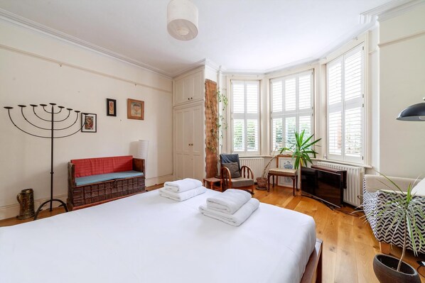 A hotel room with a large bed, a red bench, a desk with a chair, and a view of the outside through large windows. - Cosy and Spacious Home (London)