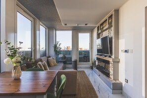 A modern living room with a dining area, large windows, a flat-screen TV, and a fireplace.