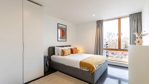 A modern bedroom with a large bed, a view of the city through a window, and a minimalist decor. - Comfortable, Modern Flat in Shoreditch (London)