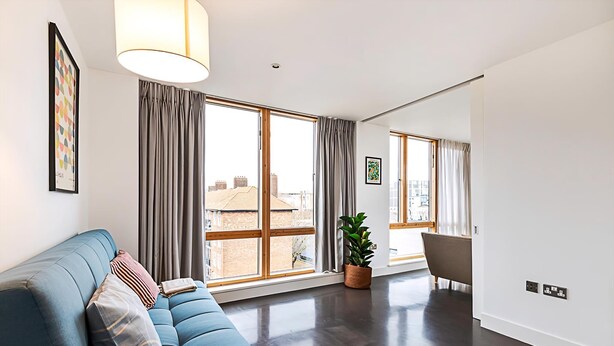 A modern living room with a blue sofa, large windows, and a potted plant. - Comfortable, Modern Flat in Shoreditch (London)