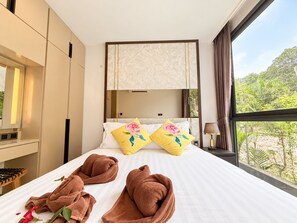 Une chambre d’hôtel avec un lit, deux oreillers ornés de motifs floraux, une table de chevet avec une lampe, et une vue sur la verdure à l’extérieur.