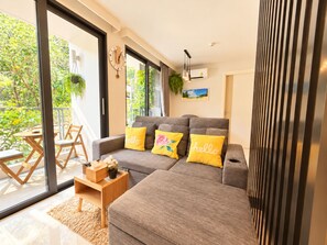 A modern living room with a grey sofa, yellow pillows, a wooden side table, and a view of greenery through large windows.