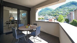 A balcony with a dining table and chairs, a view of a hillside town, and a modern interior with a lamp and a window.