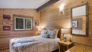 A cozy wooden cabin room with a bed, a nightstand, a table, and a window with a view of trees. - Torch Lake cabin, 4BR, Sleeps 10, Firepit, A/C (Rapid City)