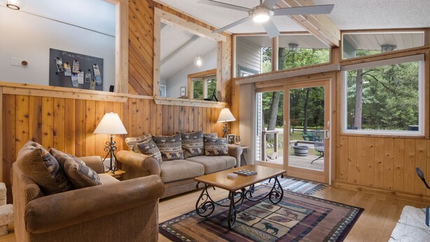 A cozy cabin interior with wooden walls, a ceiling fan, a sofa, a coffee table, and a large window offering a view of the outdoors. - Torch Lake cabin, 4BR, Sleeps 10, Firepit, A/C (Rapid City)