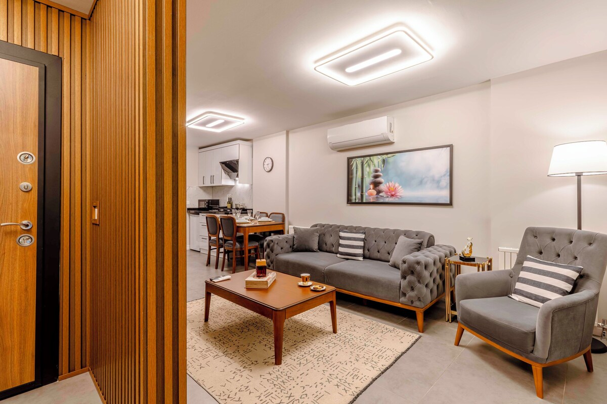 Ein modernes Wohnzimmer mit Sofa, Sessel und Couchtisch, ein Essbereich mit Stühlen und eine Küche im Hintergrund.