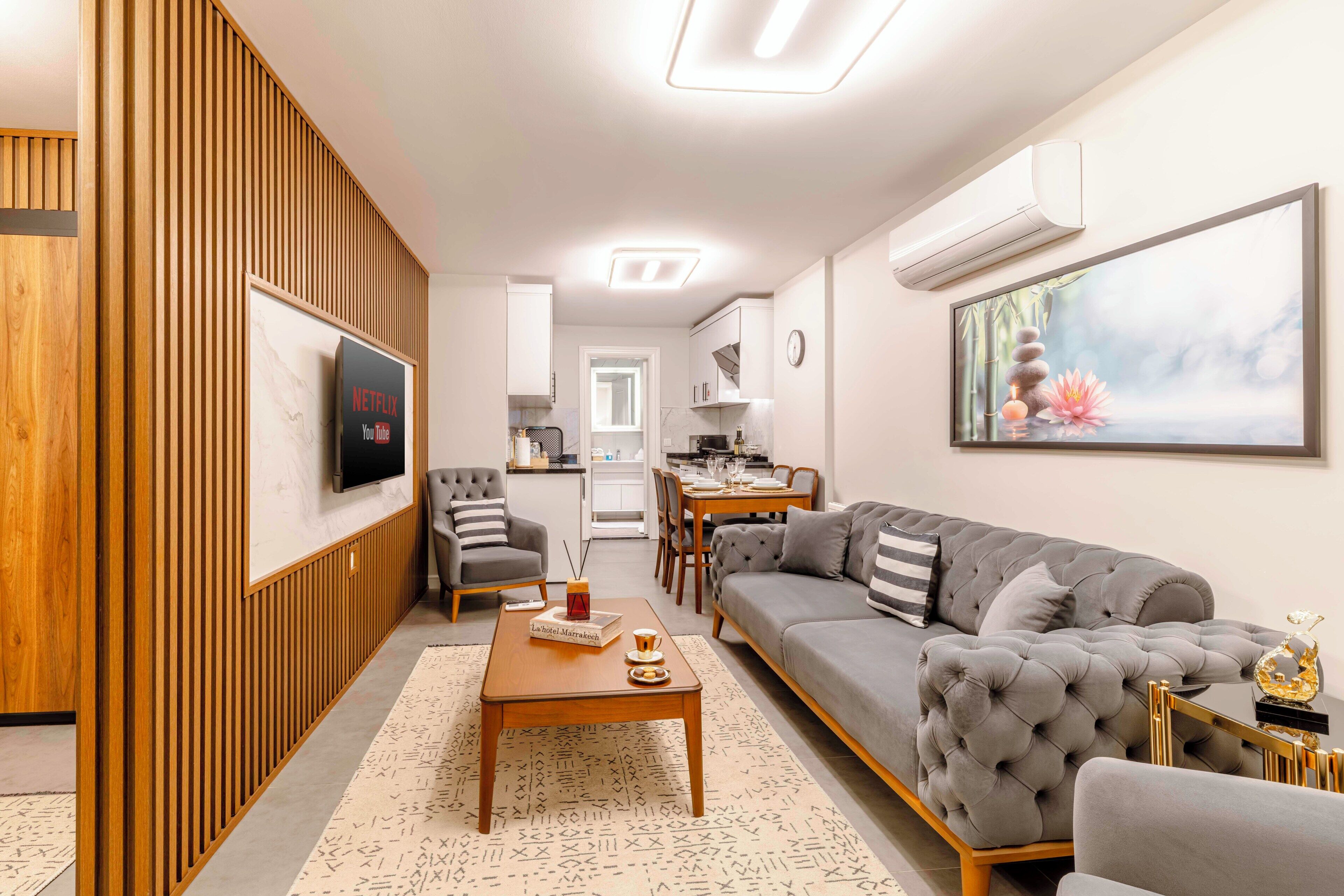 Ein modernes Wohnzimmer mit grauem Sofa, einer Wandpaneinung aus Holz, einem Flachbildfernseher und einem Essbereich mit Tisch und Stühlen.