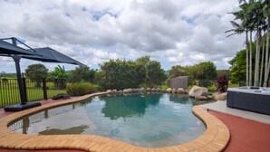 Piscina com borda curva de tijolos, rodeada por um deck de madeira e uma área coberta com um guarda-sol preto.