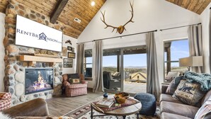 A cozy living room with a stone fireplace, a mounted deer head, and a view of the surrounding landscape. - Ozark Sunset View Log Cabin with $400 Daily Activities Included & Pickleball (Branson)