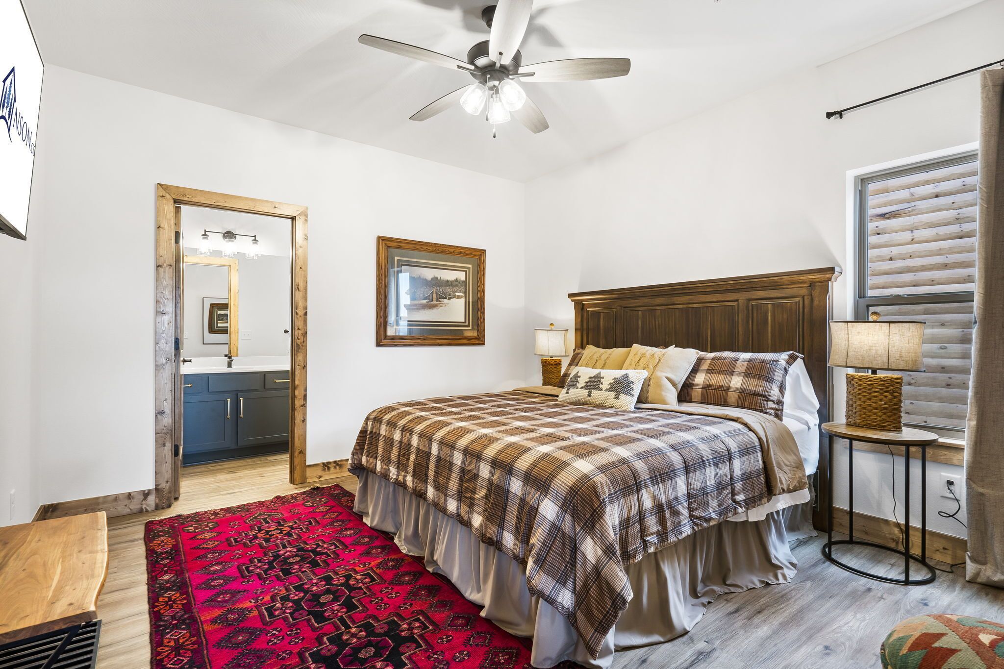 A bedroom with a bed, a ceiling fan, a window with blinds, a bedside lamp, and a red patterned rug.