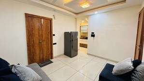 Ein modernes Wohnzimmer mit einem dunkelblauen Sofa, einer Holztür, einem schwarzen Kühlschrank und einer im Hintergrund sichtbaren Küche.