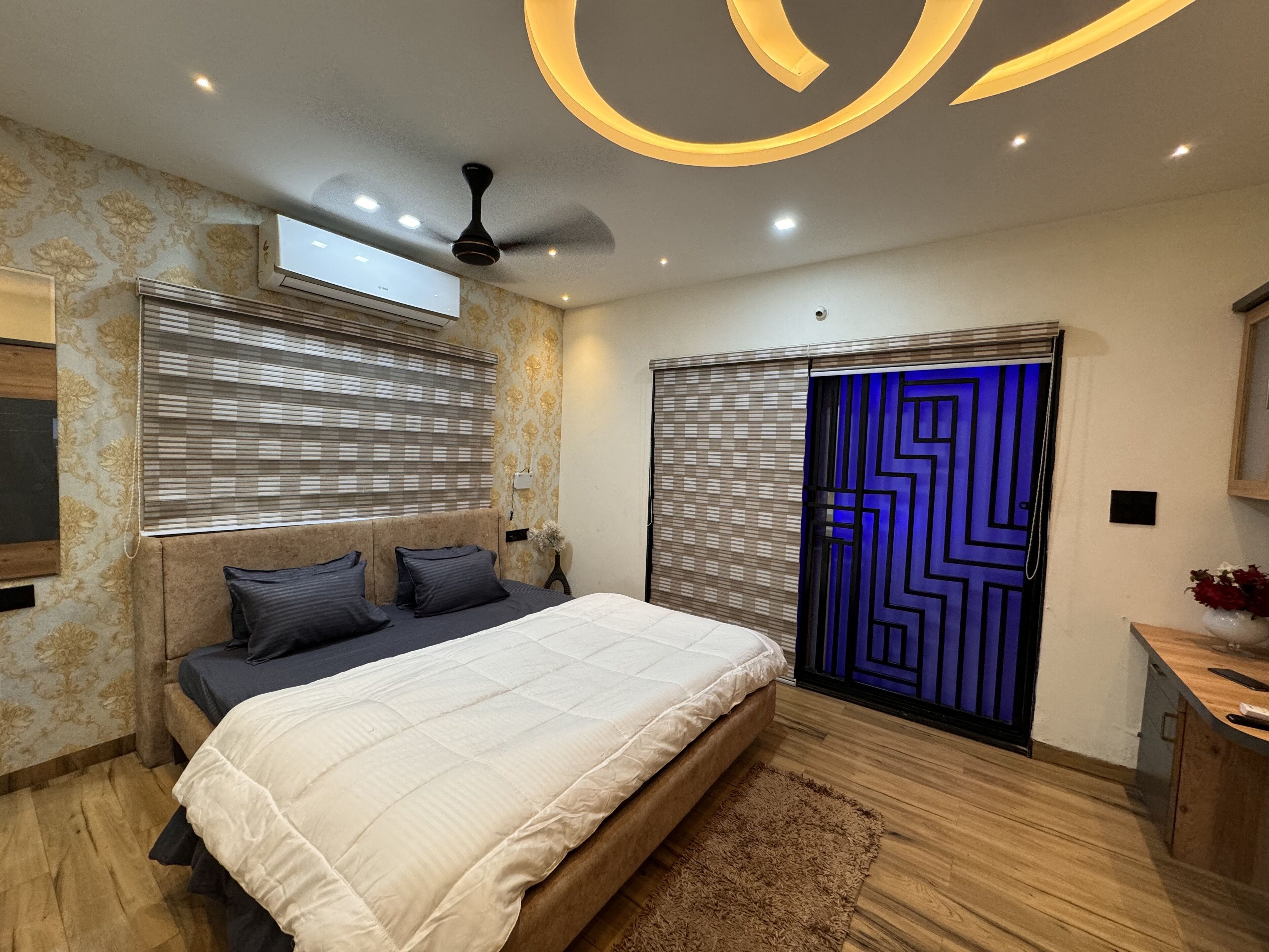 A bedroom with a bed, a ceiling fan, a window with blinds, a wall-mounted air conditioner, and a decorative wall panel.