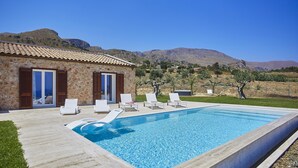 A swimming pool with lounge chairs, a stone building with shutters, and a mountainous backdrop.