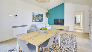 A modern living room with a wooden dining table, grey chairs, a large TV, and a painting of a coastal scene.