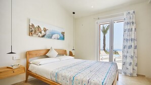 A bedroom with a wooden bed, a nightstand, a painting of a coastal scene, and a view of the sea through a glass door.