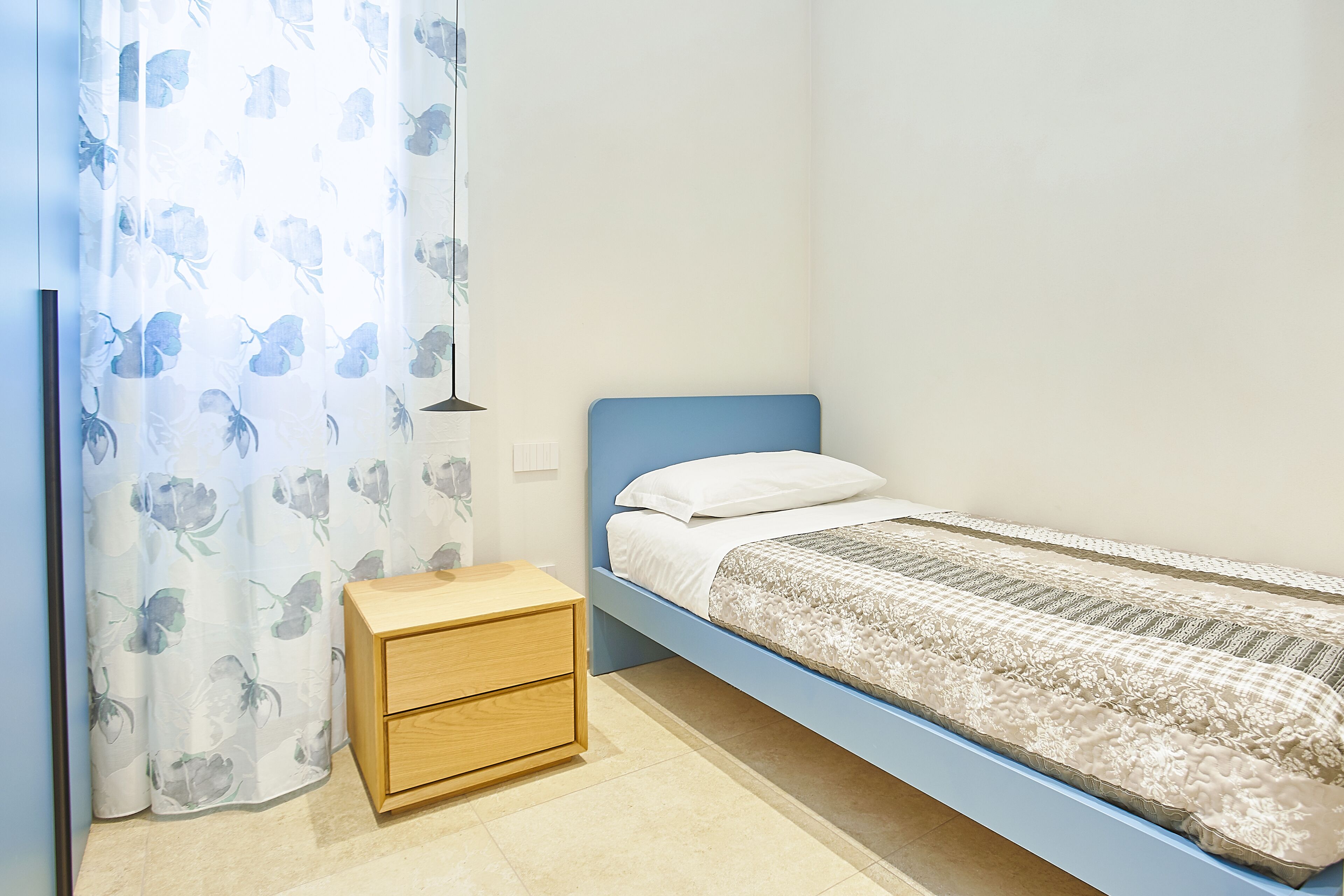 A single bed with a blue headboard, a wooden nightstand with drawers, and a floral-patterned curtain.