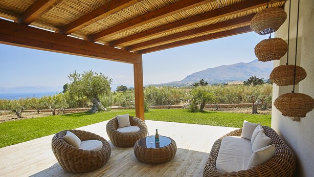 A covered outdoor seating area with wicker furniture and a view of a vineyard and mountains.