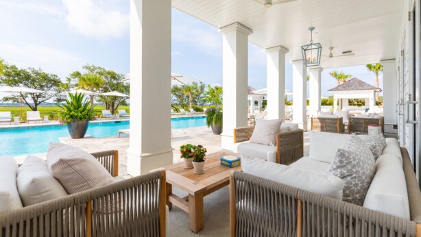 A covered outdoor seating area with wicker furniture, a pool in the background, and a view of the ocean.