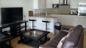 A modern living room with a flat-screen TV, a glass coffee table, a leather sofa, and a kitchen area with white cabinets and stainless steel appliances. - Britain in the Sun at Luxurious Ocean Village (Gibraltar)