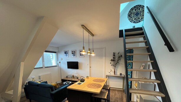Un moderno loft con una scala in legno, una zona pranzo con tavolo in legno, un divano blu e un televisore montato a parete.