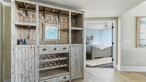 A rustic wooden cabinet with wine glasses and a framed picture, a bed with a headboard in the background, and a hallway leading to another room. - Family-Friendly Two Bedroom Unit with Phenomenal Gulf and Bay Views. Complimentary Boat Slip! (Orange Beach)