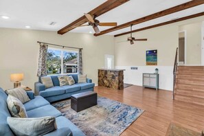 A spacious living room with a blue sofa, a coffee table, and a wooden floor. There is a ceiling fan, a window with curtains, and a kitchen area in the background. - Nokomis Beach Getaway with Heated Pool (Nokomis)