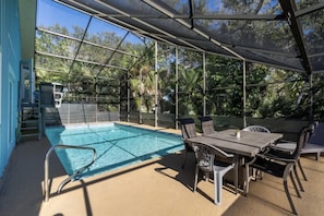 Una piscina exterior con cerramiento de vidrio, rodeada de sillas y una mesa, con árboles al fondo.