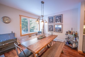 Un coin repas comprenant une table en bois, des chaises, un lustre et une fenêtre donnant sur des arbres.