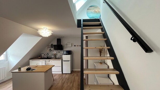 A modern kitchen with a central island, a staircase with wooden steps, and a view of the outdoors through a skylight. - Studio Scandinave - Duplex - Hyper Centre (Lorient)