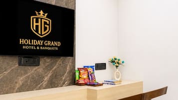 A hotel reception desk with a mounted TV displaying the hotel's logo, a power outlet, snacks, a vase with flowers, and a wooden chair with a cushion.