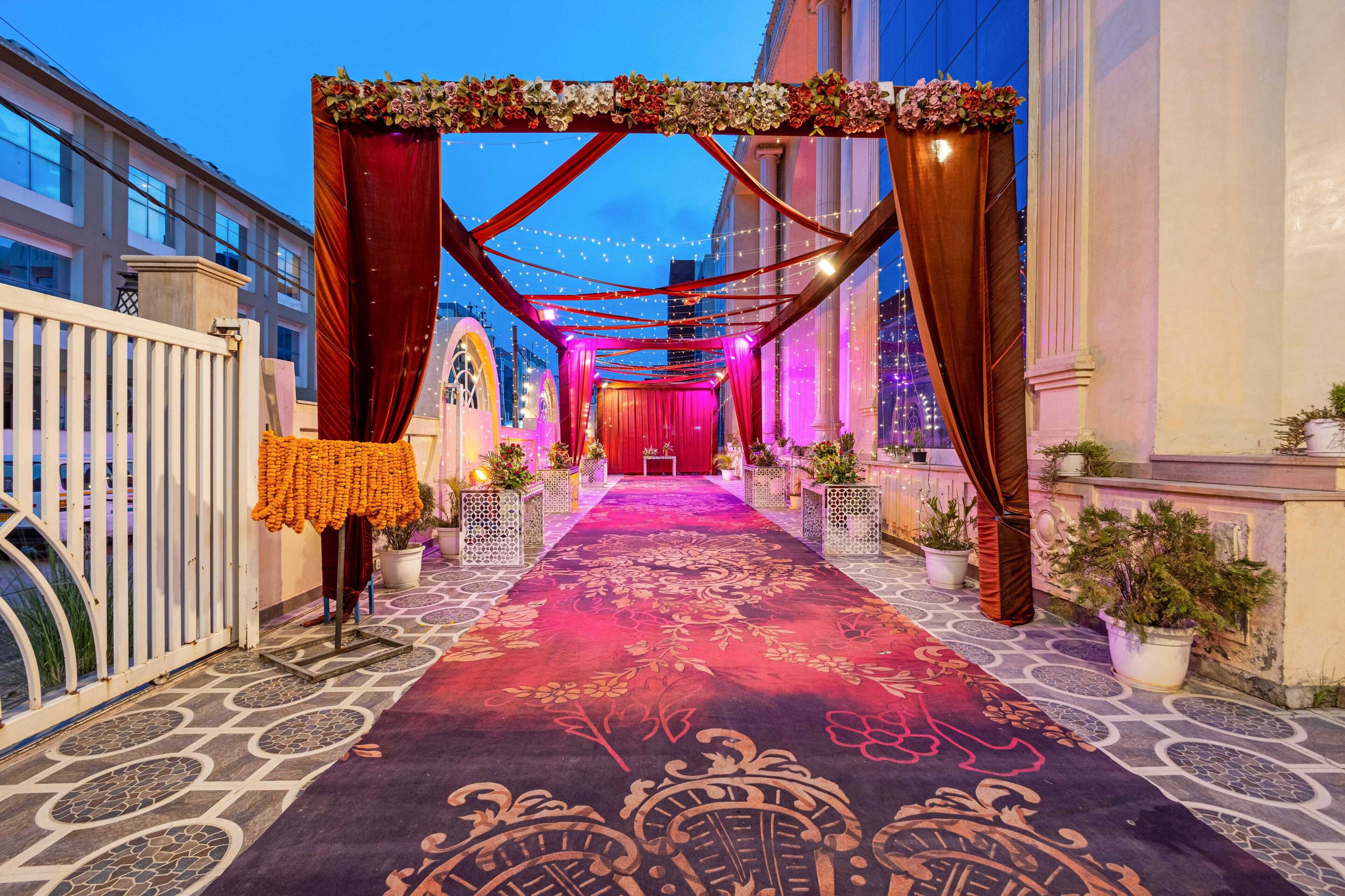 Un pasillo exterior con alfombra roja, arcos decorativos e iluminacion con guirnaldas.